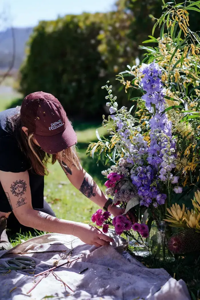 Christina kneeling on the grass arranging delphiniums and wattle into a vase
