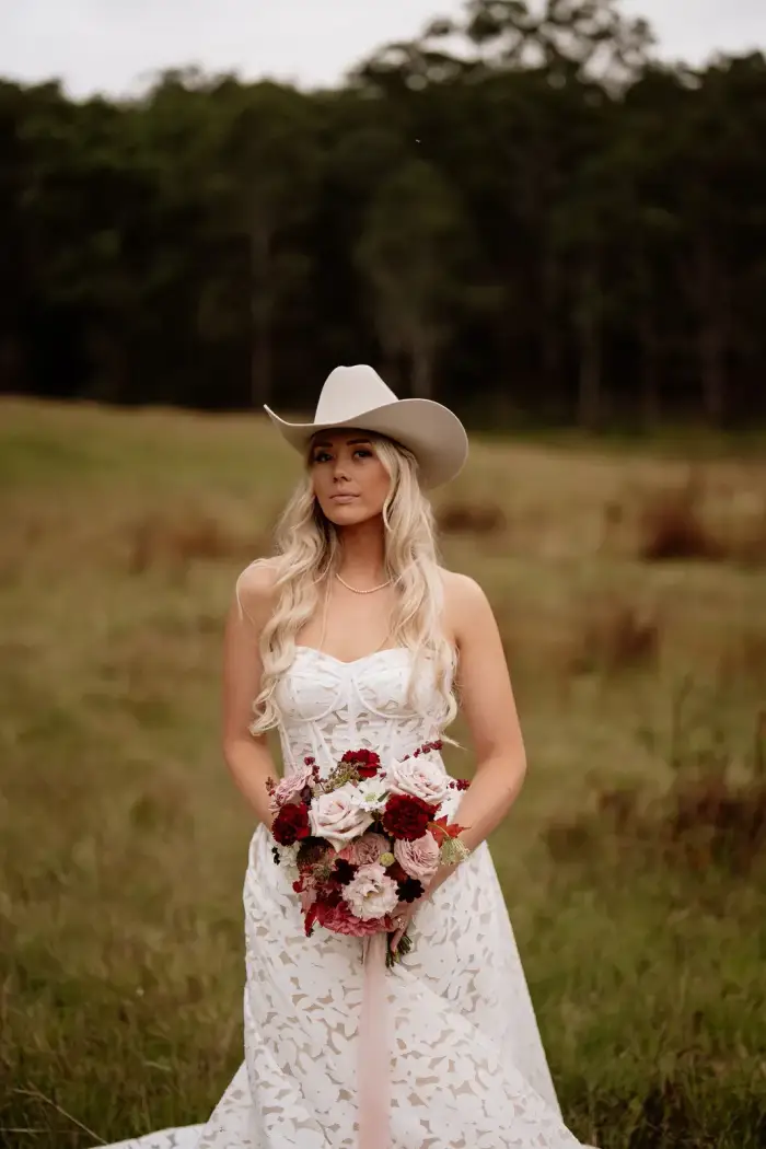 Bride in a white hat holding a burgundy and blush bouquet