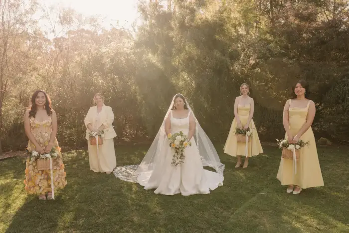 Bride and her bridesmaids in yellow holding white and yellow bouquets on a sunlit lawn