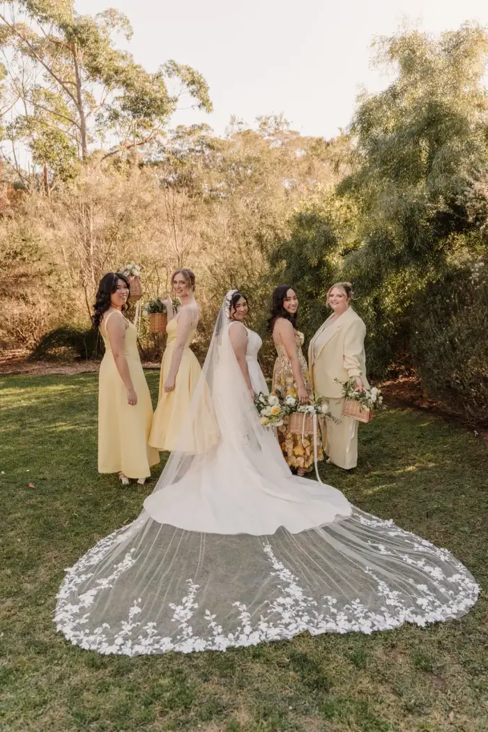 Bride from behind with her lace veil spread across the grass, bridesmaids either side