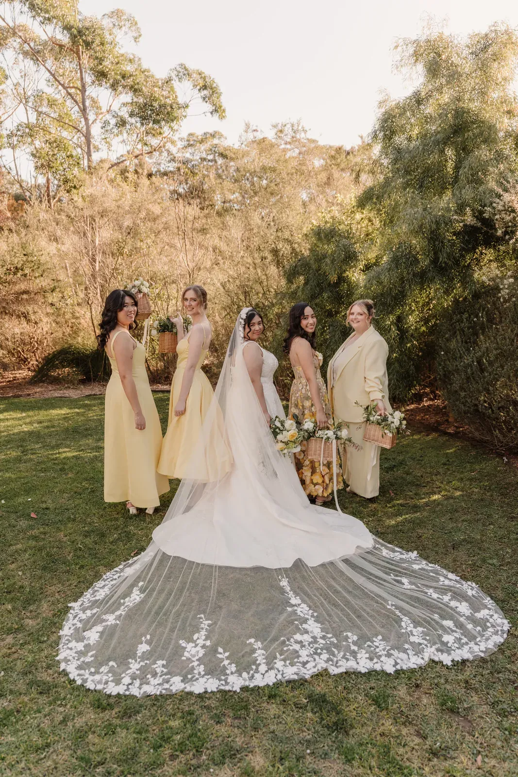 Bride from behind with her lace veil spread across the grass, bridesmaids either side