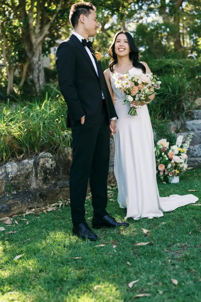 Bride and groom smiling at each other beside a stone garden wall