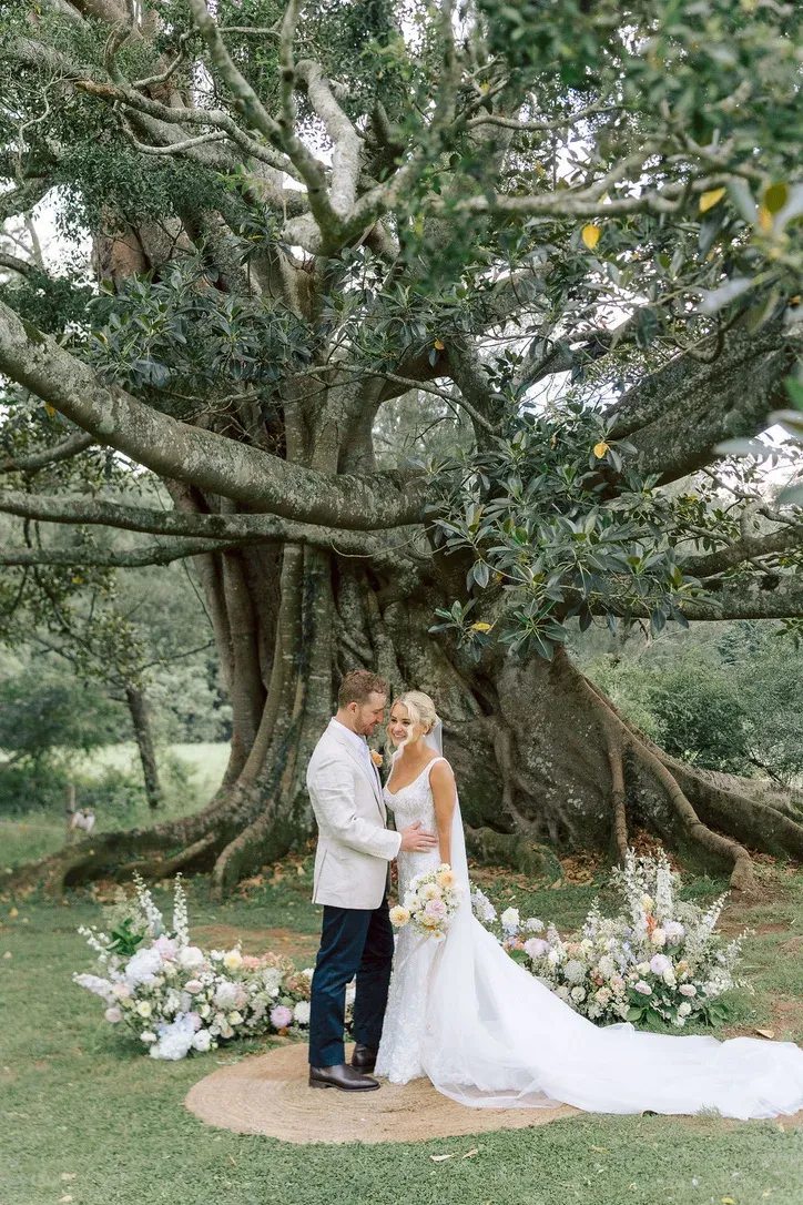 Bride and groom embracing beneath a heritage fig tree, styled with a pastel floral ceremony arrangement