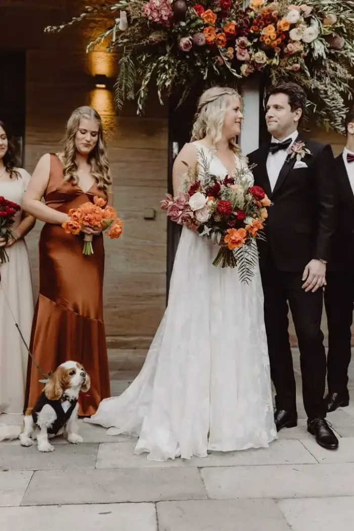 Bridal party outside a venue entrance at night, with bouquets in warm autumn tones and the couple's dog